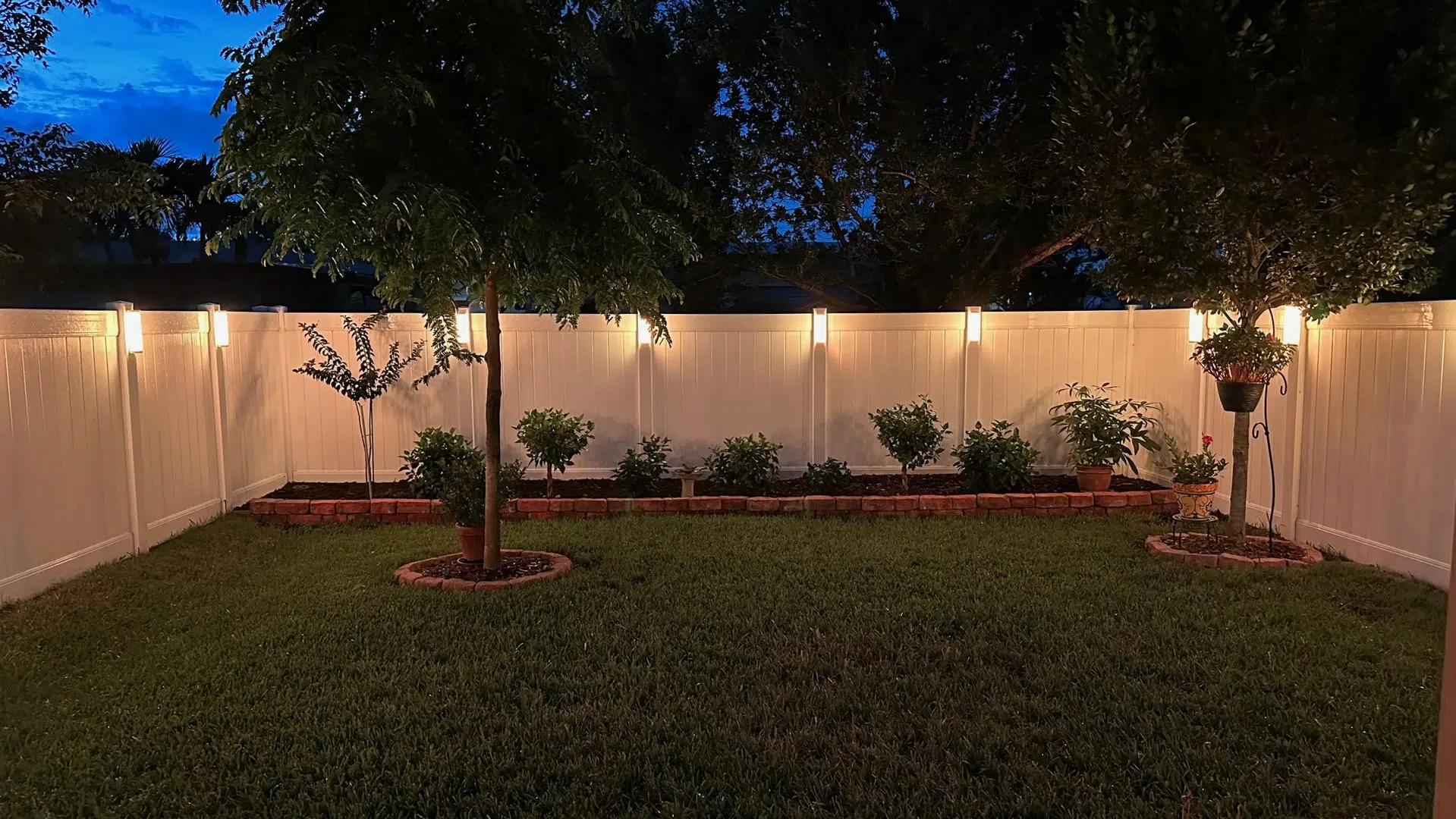 Soft white lighting along a backyard fence and tree line in an Odessa home at sunset.